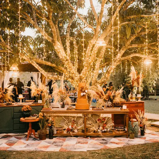 Rustic wedding dessert table beneath fairy-light-draped tree with tiered cake
