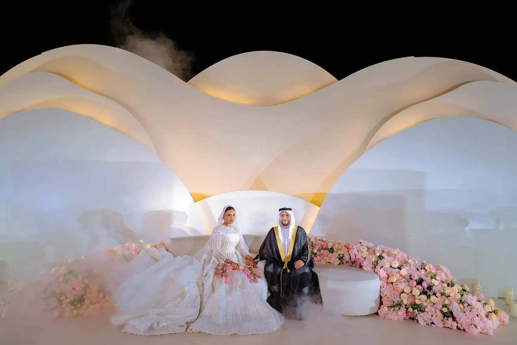 Bride and groom seated at sculptural white stage surrounded by pink roses in Dubai desert